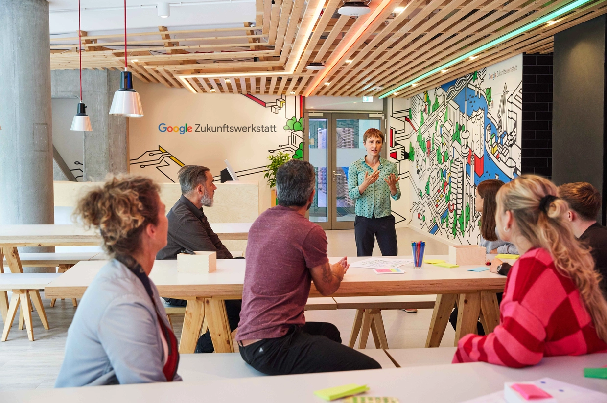 In einem hellen Seminarraum der Google Zukunftswerkstatt sitzt eine Gruppe von Menschen an Tischen und hört einer Trainerin zu, die vor ihnen spricht. An der Wand sind das Logo der Google Zukunftswerkstatt und eine großflächige Illustration zu sehen.