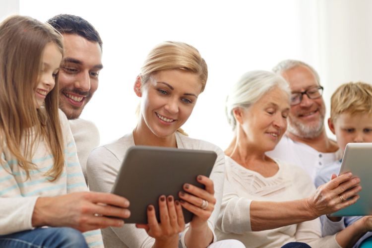 Mehrere Personen unterschiedlichen Alters sitzen gemeinsam auf einem hellen Sofa und betrachten lächelnd zwei Tablets.